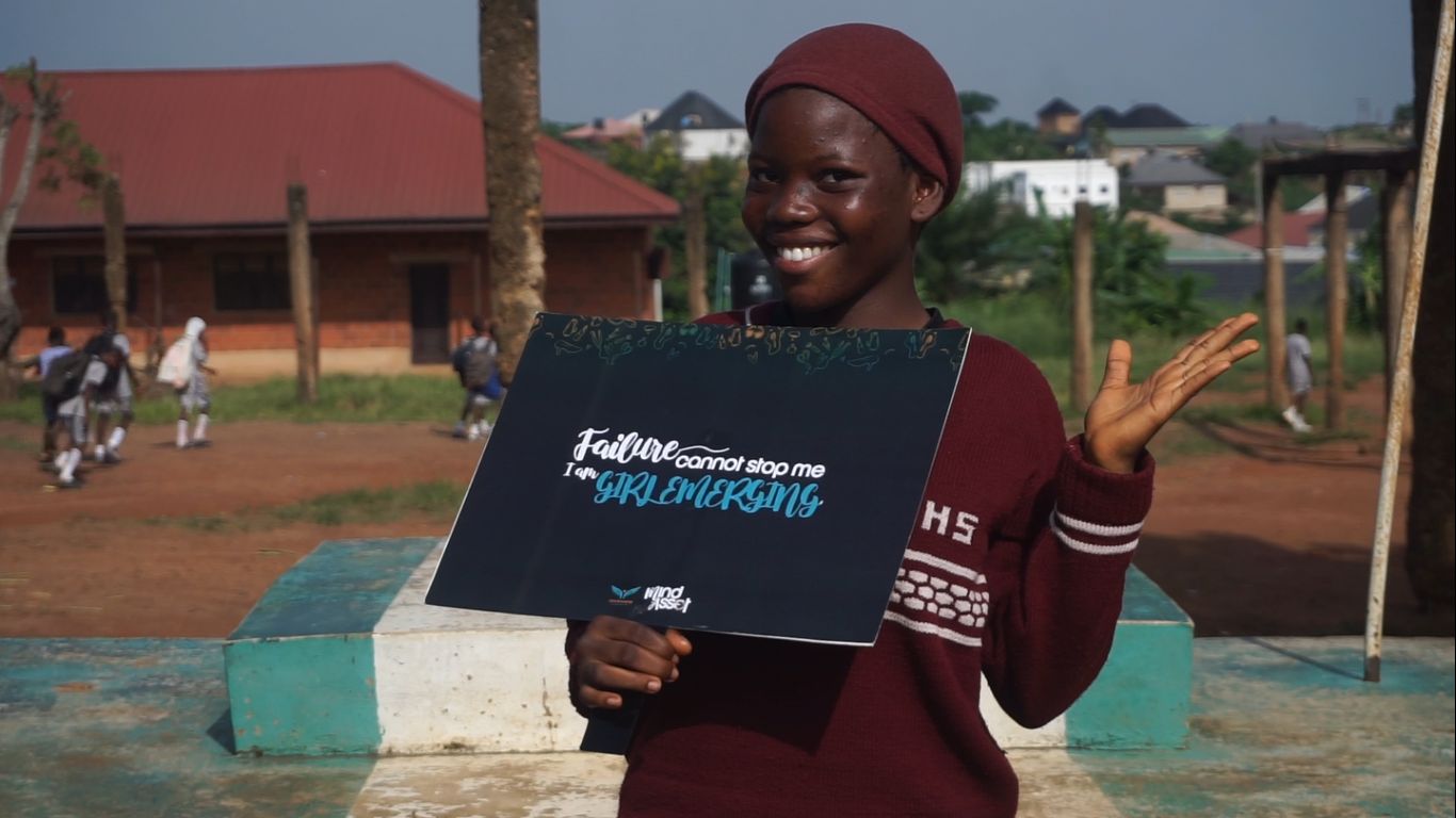 A girl smiling while holding a Girls Emerging sign outside her school